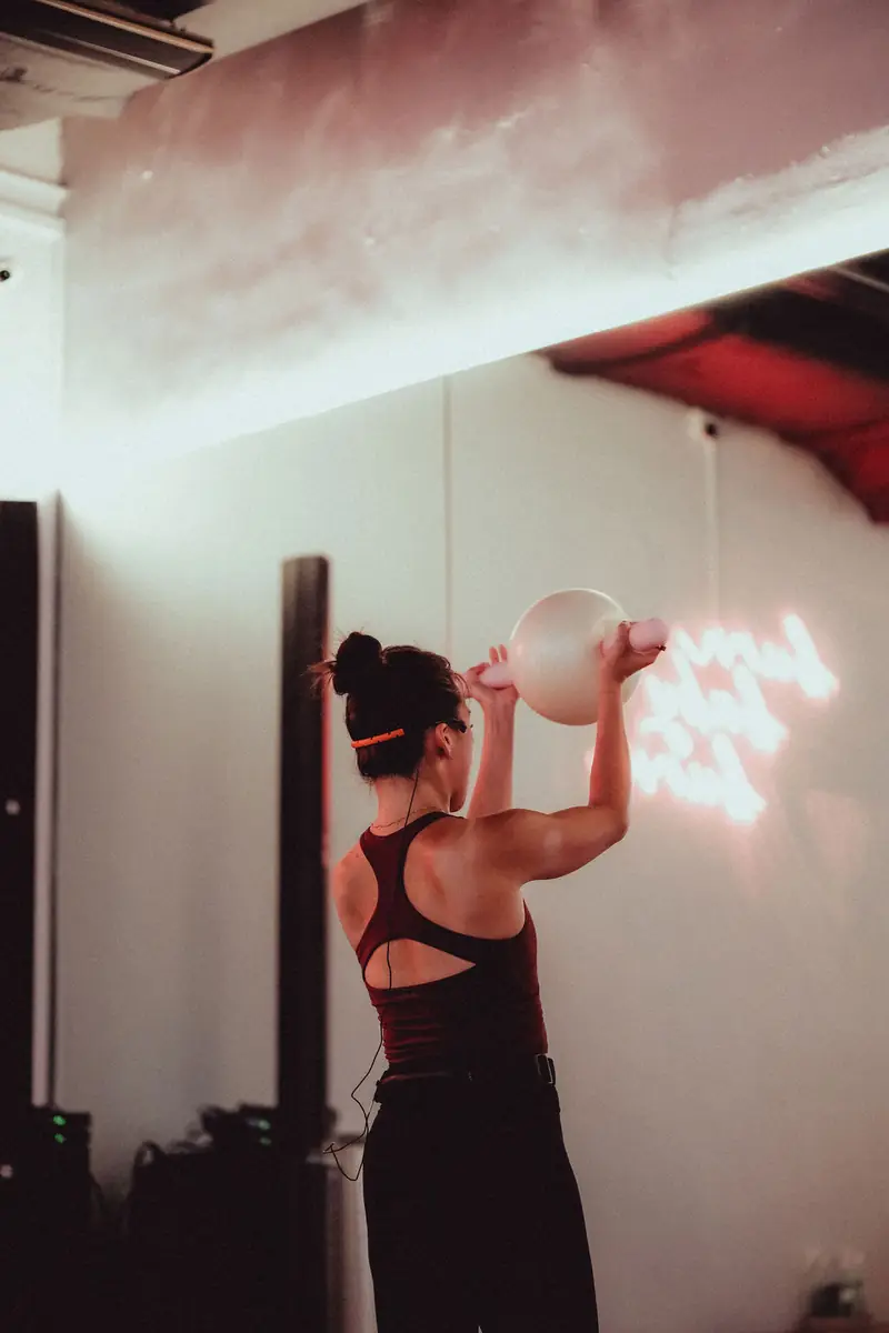 A woman holding a pilates ball overhead by a pink neon sign