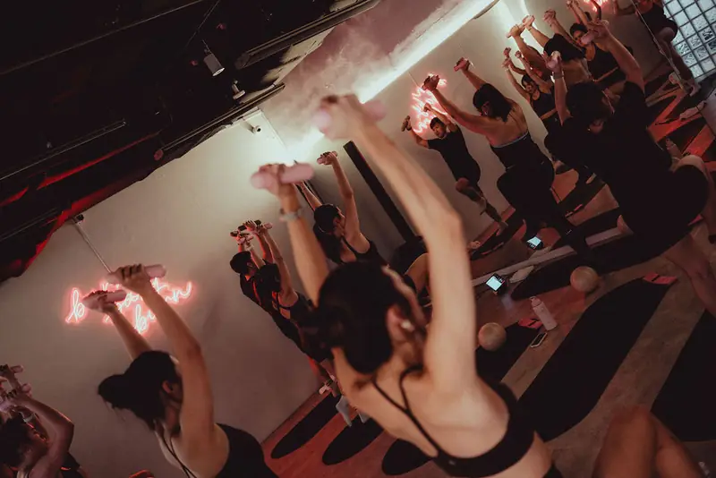 A Hot Mat Pilates class with arms up under pink neon, seen from above