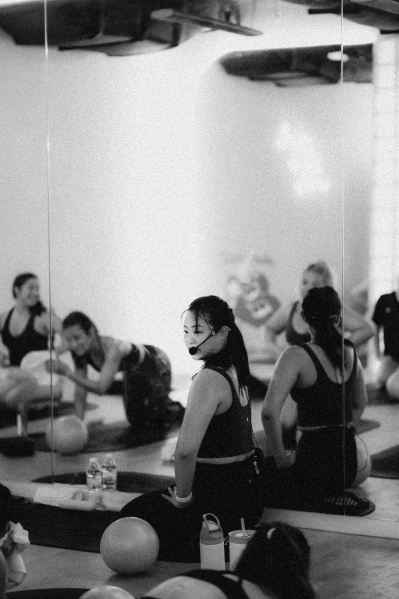 Black and white photo of an instructor with a headset sitting with her class on their mats