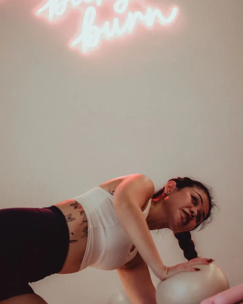 A woman on her mat with a pilates ball under a pink neon sign